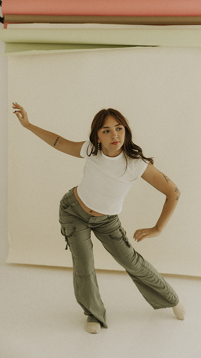 Pointe toe with an exaggerated bend with alternate arm extended by a brunette dancer in a white tee and olive pants