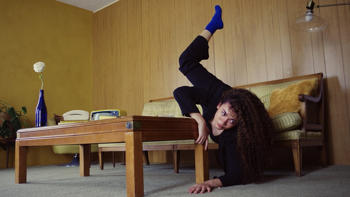 Jordan in black dance attire and blue socks in an exaggerated falling dance pose in a retro living room set