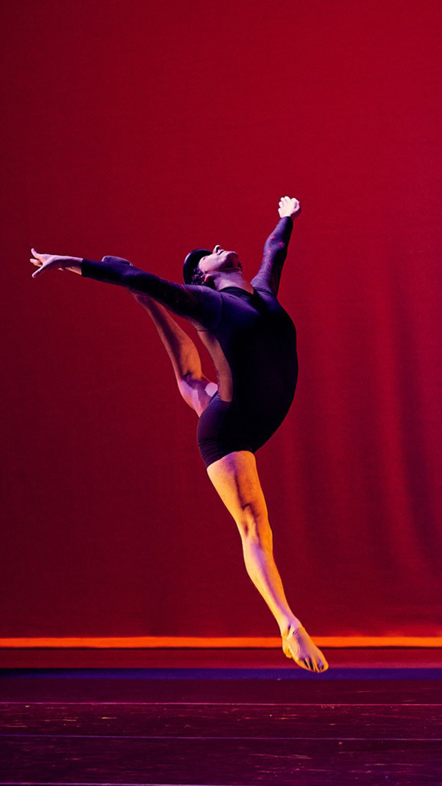 Scarlet red backdrop and lighting illuminates the dance leap done by Jonathan Montoya Gerardo in a royal purple velvet dance suit